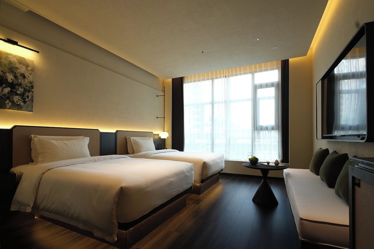 A hotel room with two beds, a sofa, a small table, and a large window with curtains.
