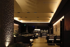 A hotel corridor with a modern design, featuring a central seating area with tables and chairs, a textured wall on the left, and a ceiling with recessed lighting. - YOMANJO (Guangzhou)