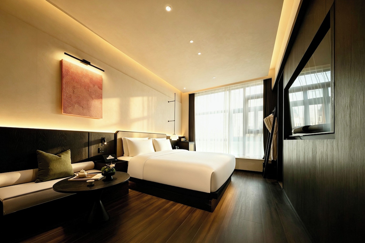 A modern hotel room with a large bed, a sofa, a small table, and a large window.