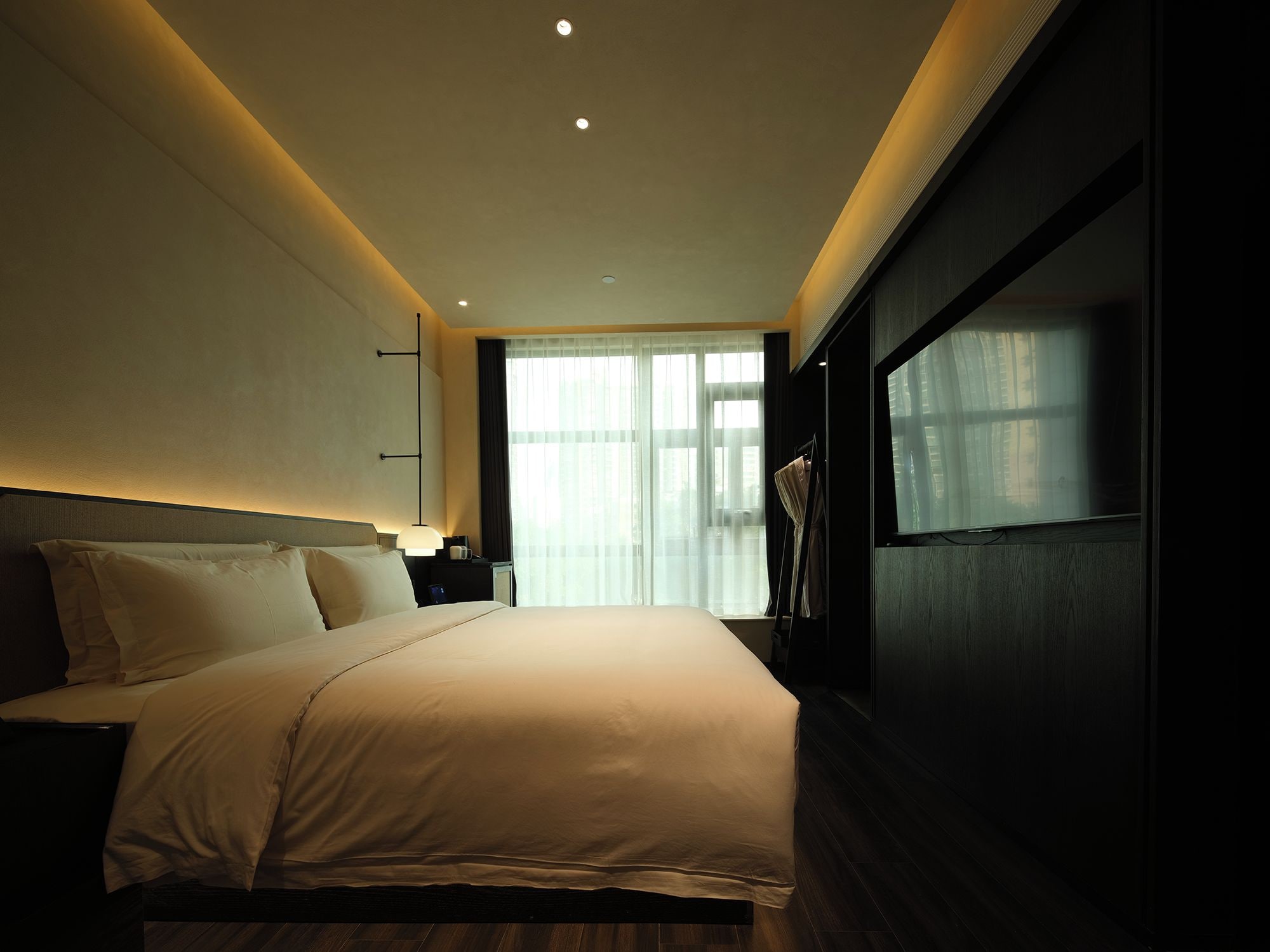 Una habitación de hotel moderna con una cama grande, una mesita de noche y un amplio ventanal con cortinas.