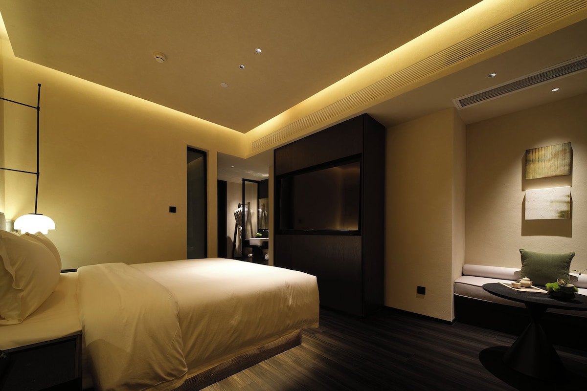 A modern hotel room with a bed, bedside table, and a seating area with a table and cushions.