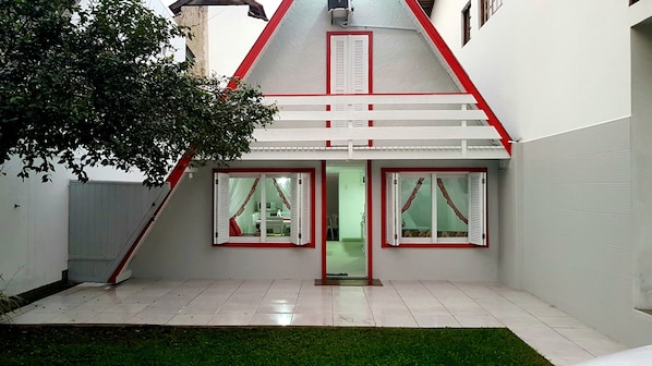 Uma casa com telhado inclinado, paredes externas brancas e um pequeno jardim frontal com gramado.