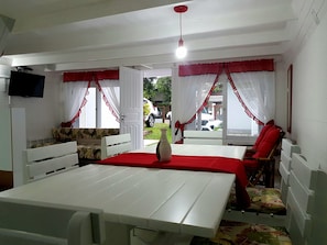 Quarto com uma mesa branca, duas cadeiras vermelhas, um sofá com estampa floral e uma televisão fixada na parede.