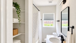 Un baño moderno con un lavamanos blanco, un grifo negro y un espejo grande. Hay una ducha con cortina blanca, una ventana con persianas y una planta colgando del techo.
