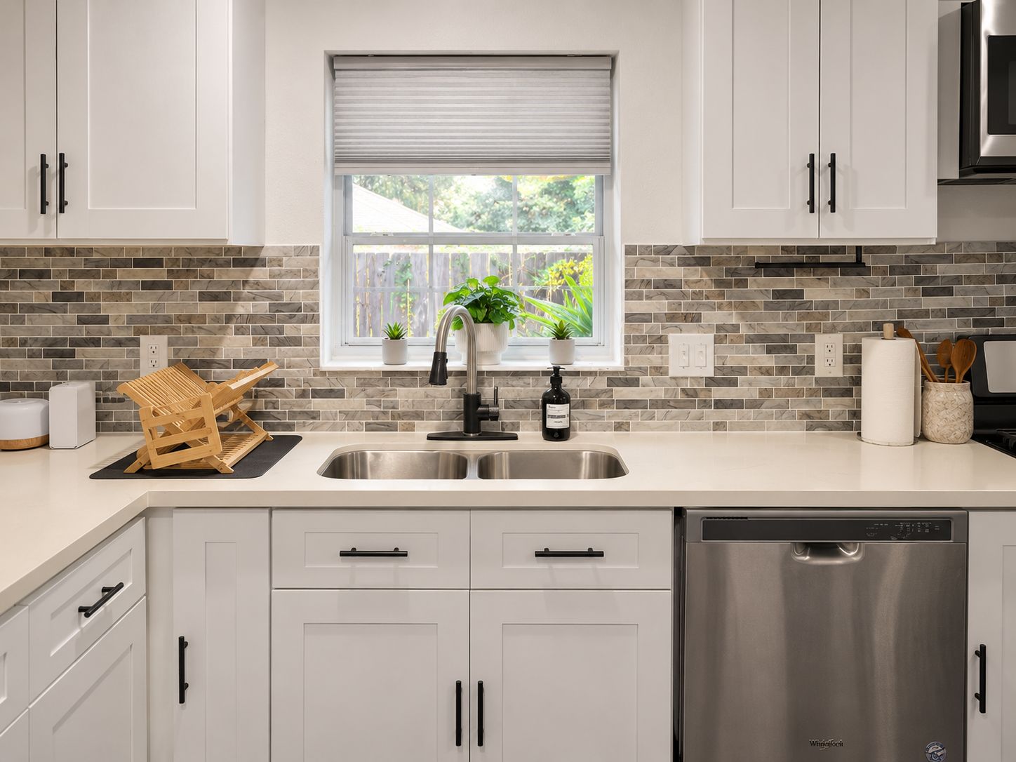 A modern kitchen with a stainless steel sink, a built-in dishwasher, and a window with a view of greenery.