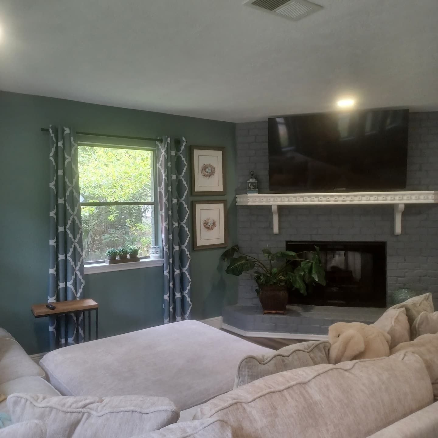 A living room with a fireplace, a sofa, a TV mounted above the fireplace, and a window with curtains.