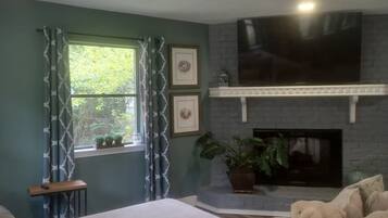A living room with a fireplace, a sofa, a TV mounted above the fireplace, and a window with curtains.