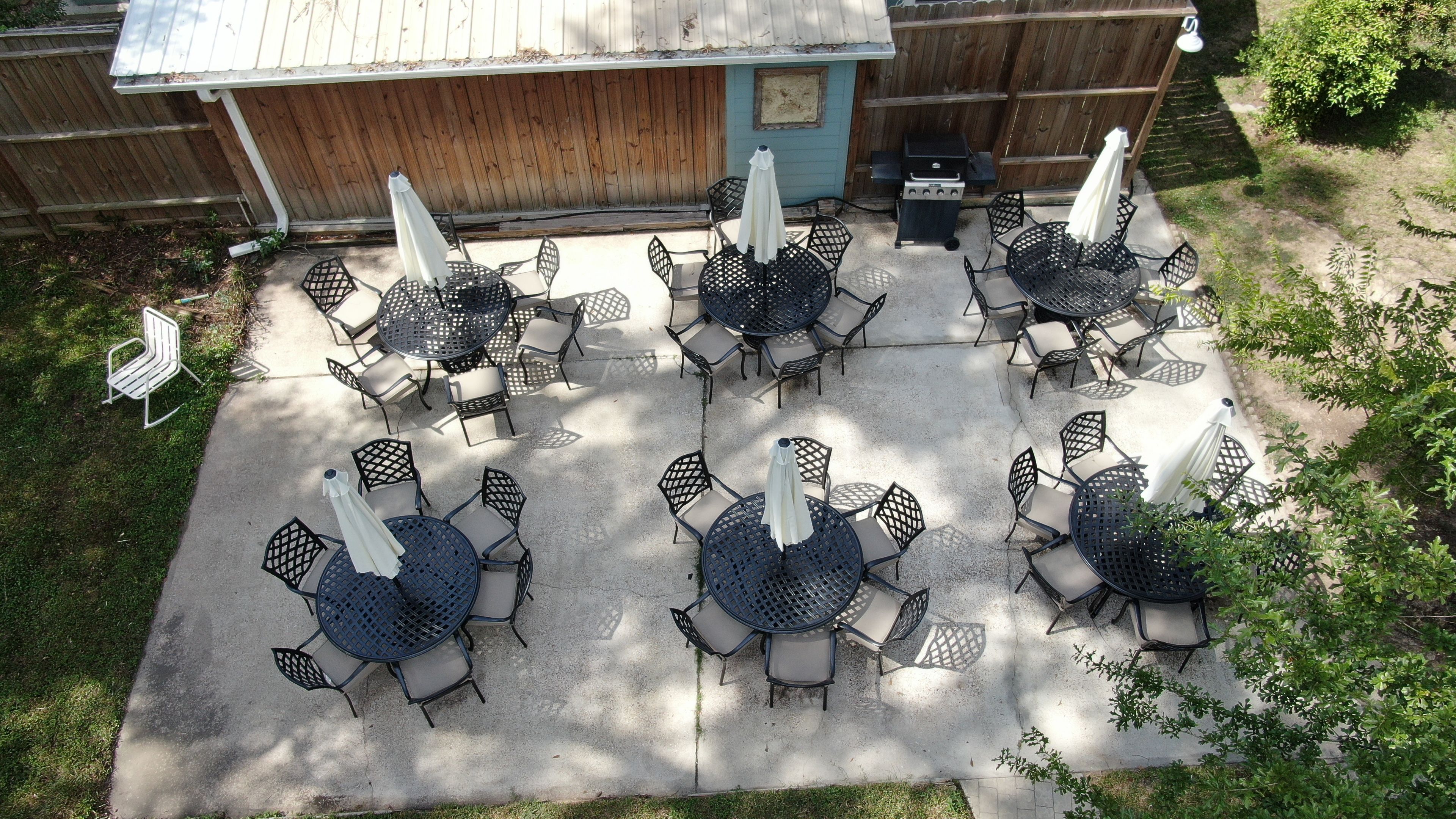 An outdoor patio with multiple round tables and chairs, a barbecue grill, and a wooden fence in the background.