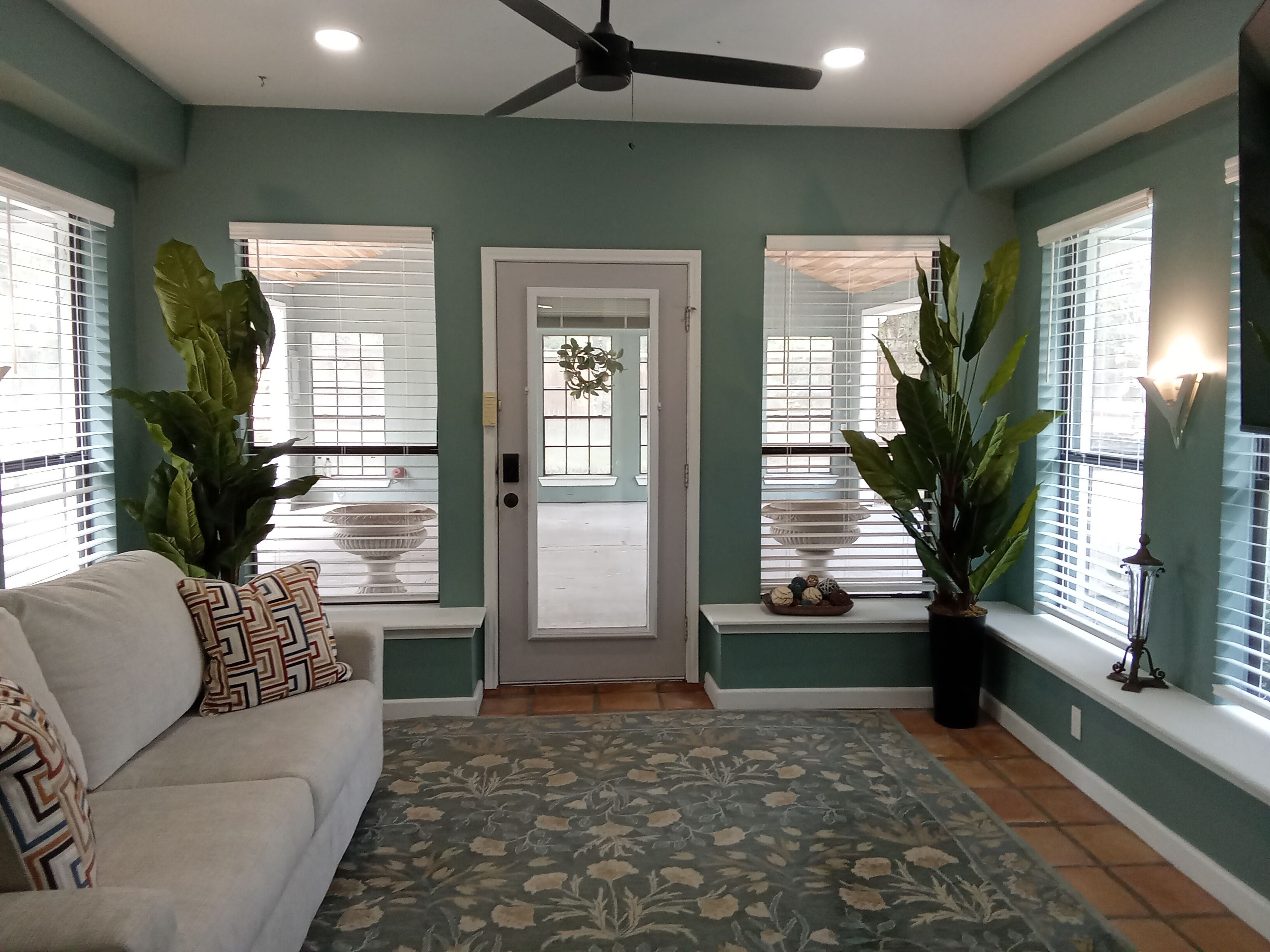 A living room with a ceiling fan, a sofa with patterned cushions, a decorative bowl on a windowsill, and a large potted plant.
