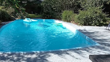 Een zwembad met een helderblauw wateroppervlak, omgeven door bomen en een overdekt terras met een ligstoel.