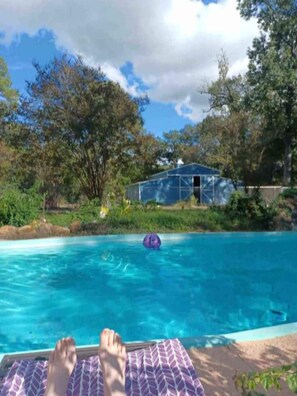 A swimming pool with a person relaxing on a lounge chair, surrounded by trees and a greenhouse structure.