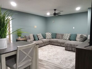 A living room with a sectional sofa, a dining table with chairs, a ceiling fan, and a potted plant.