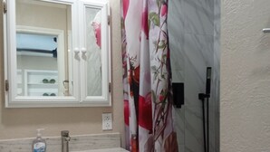 Ein Badezimmer mit weißem Waschtisch, einem Spiegel, einer Duschvorhang mit Blumenmuster und einer Handbrause.