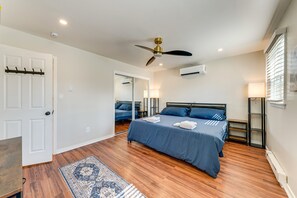 A bedroom with a bed, bedside tables, a ceiling fan, an air conditioning unit, and a window with blinds. - The Buttonwood Retreat Game Room Fire Pit (Tobyhanna)