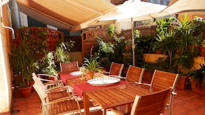 Une terrasse extérieure avec une table et des chaises en bois, entourée de plantes en pot et surmontée d’un toit de chaume.
