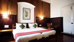 Una cama doble con una alfombra roja, dos mesitas de noche, un buró, un cuadro en la pared y una puerta blanca.