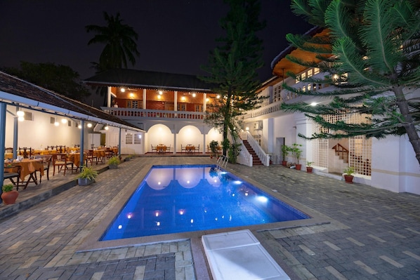 Una piscina con solárium de baldosas, rodeada de mobiliario exterior y un edificio con varios niveles y balcones.
