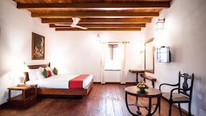 Una habitación de hotel con techo de madera, una cama con manta roja, una mesita de noche con lámpara, una mesa redonda con flores y una silla.
