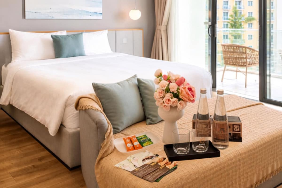 Quarto de hotel com cama, mesa de cabeceira com um vaso de flores, bandeja com garrafas e copos, e vista da cidade pela janela.