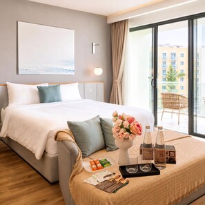 Quarto de hotel com cama, mesa de cabeceira com um vaso de flores, bandeja com garrafas e copos, e vista da cidade pela janela.
