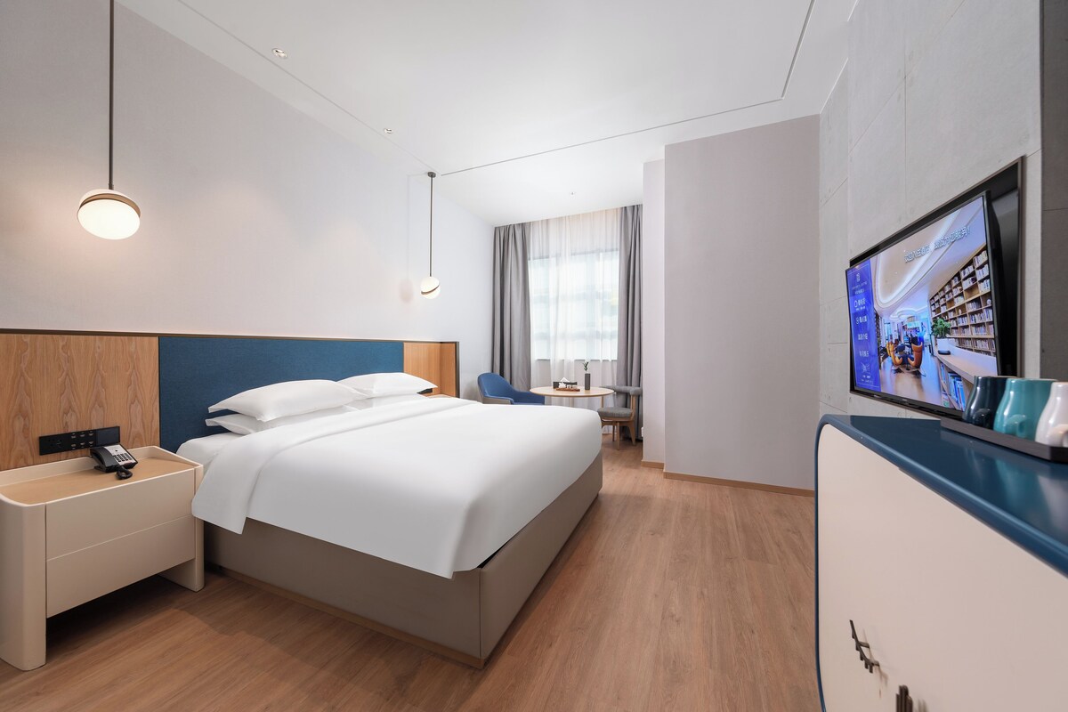Habitación de hotel moderna con una cama grande, mesitas de noche, un escritorio, una silla, un televisor y una ventana con cortinas.