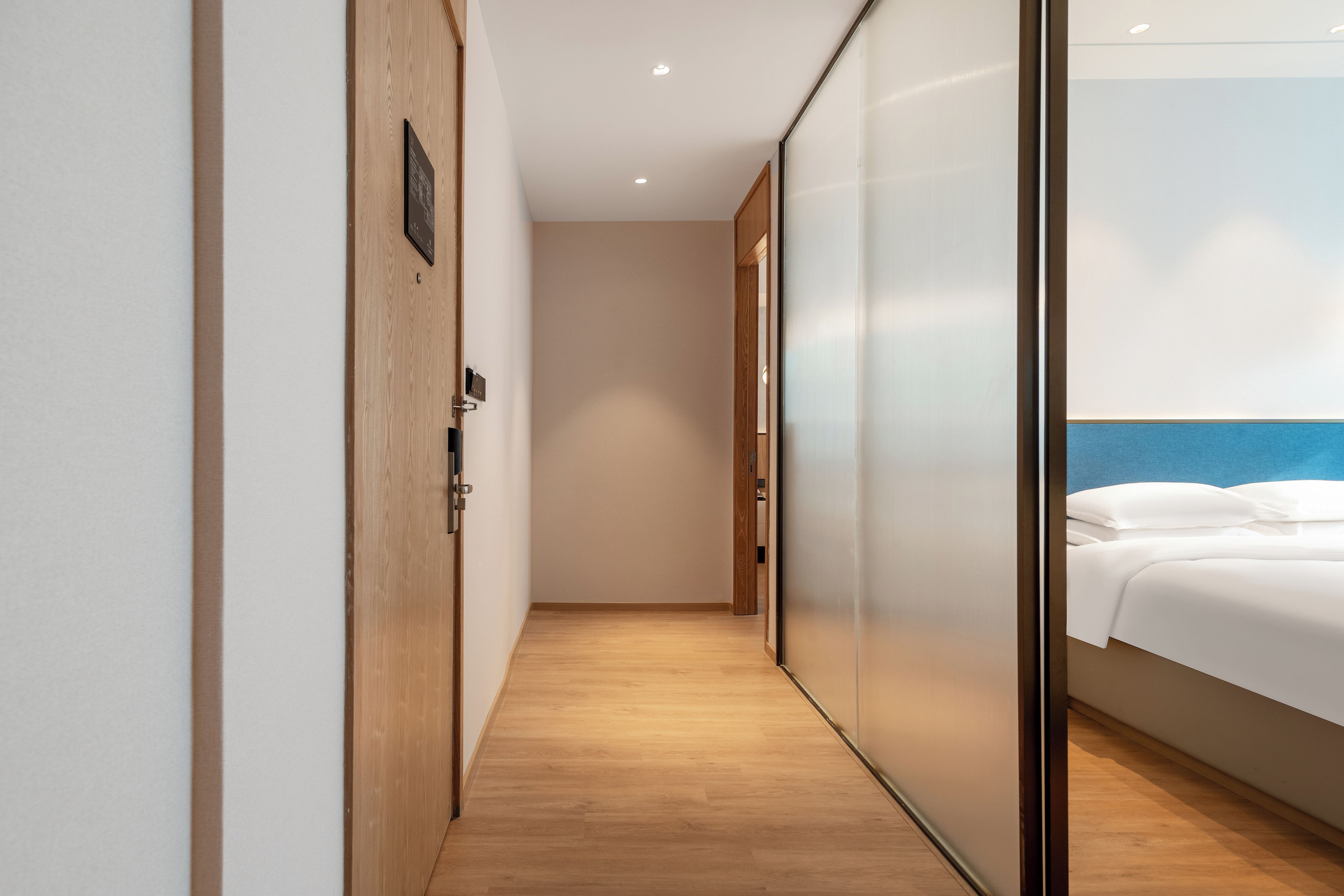 A hotel corridor with a wooden floor, a sliding door leading to a room with a bed and a blue headboard.