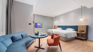 Une chambre d’hôtel avec un canapé bleu, une table ronde, une chaise orange, un lit avec une literie blanche, une table de chevet avec un téléphone et un téléviseur fixé au mur.
