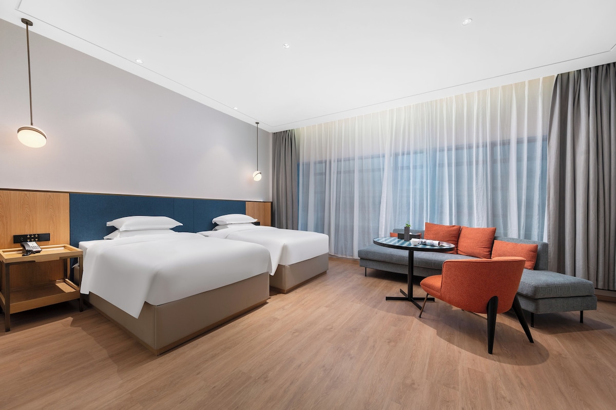 Habitación de hotel con dos camas, un sofá, una mesita y una silla. Se observa un buró con un teléfono y una lámpara.