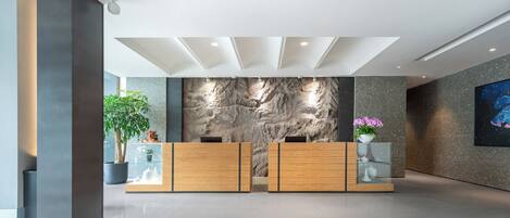 A reception area with a modern design, featuring a reception desk, a wall with a textured stone-like pattern, and a potted plant.