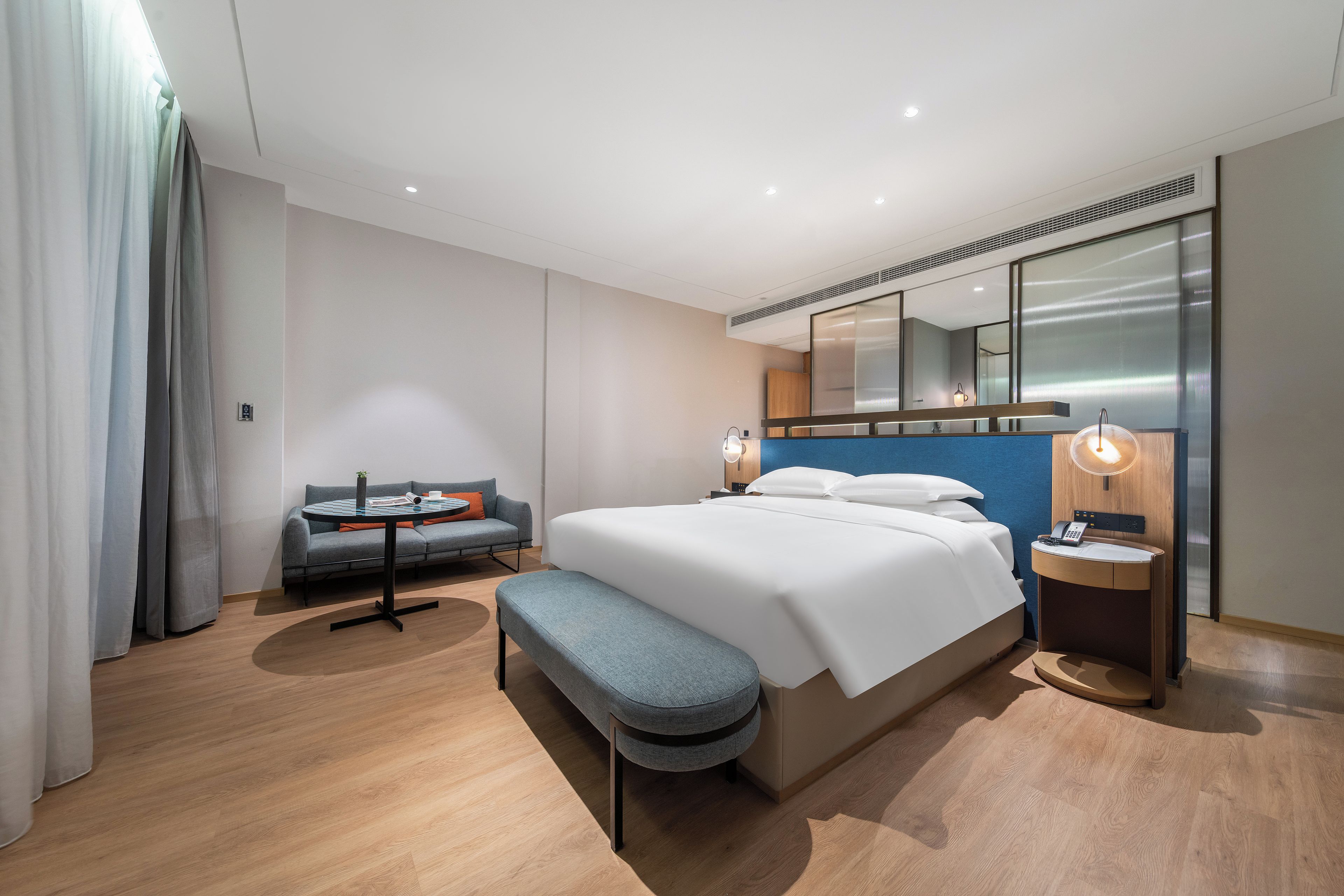 A modern hotel room with a large bed, a seating area with a sofa and a small table, a bench, a bedside table with a lamp, and a large mirror above the bed.