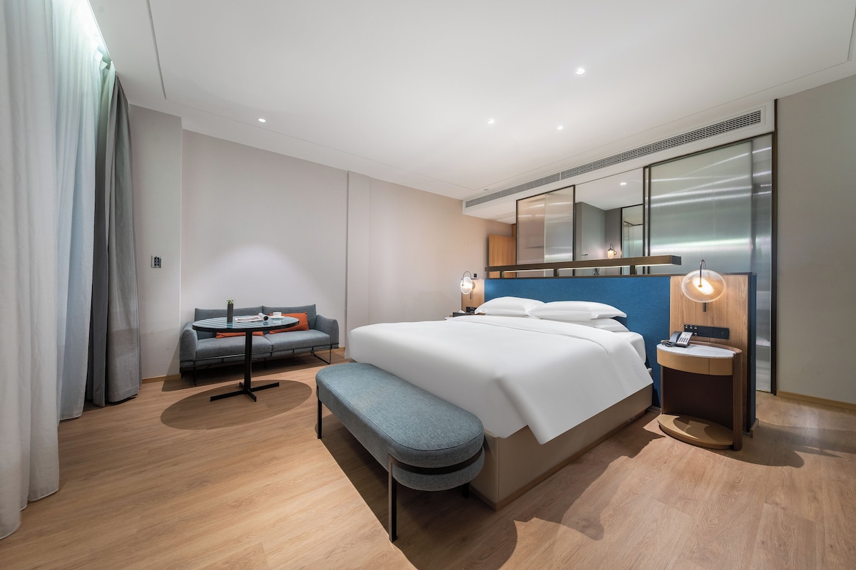 Una habitación de hotel moderna con una cama grande, una zona de estar con un sofá y una mesita, un banco, una mesita de noche con lámpara y un espejo grande sobre la cama.