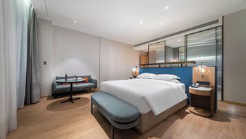 A modern hotel room with a large bed, a seating area with a sofa and a small table, a bench, a bedside table with a lamp, and a large mirror above the bed.