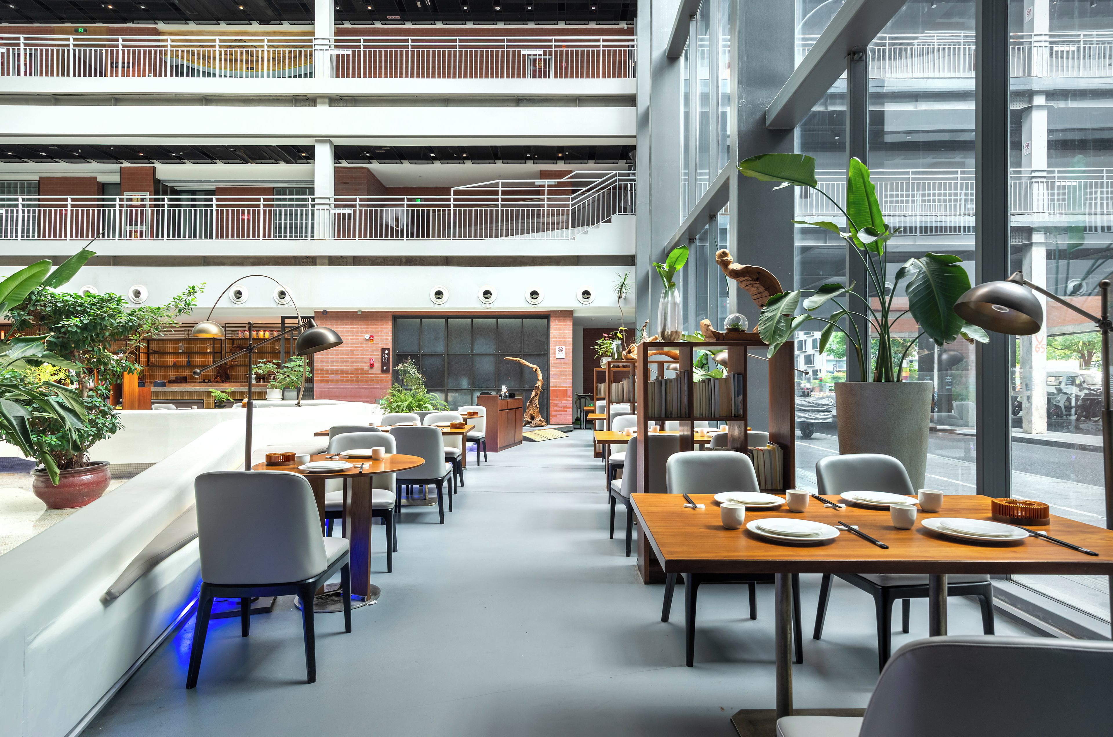 A modern indoor space with multiple dining tables and chairs, large windows, and indoor plants.