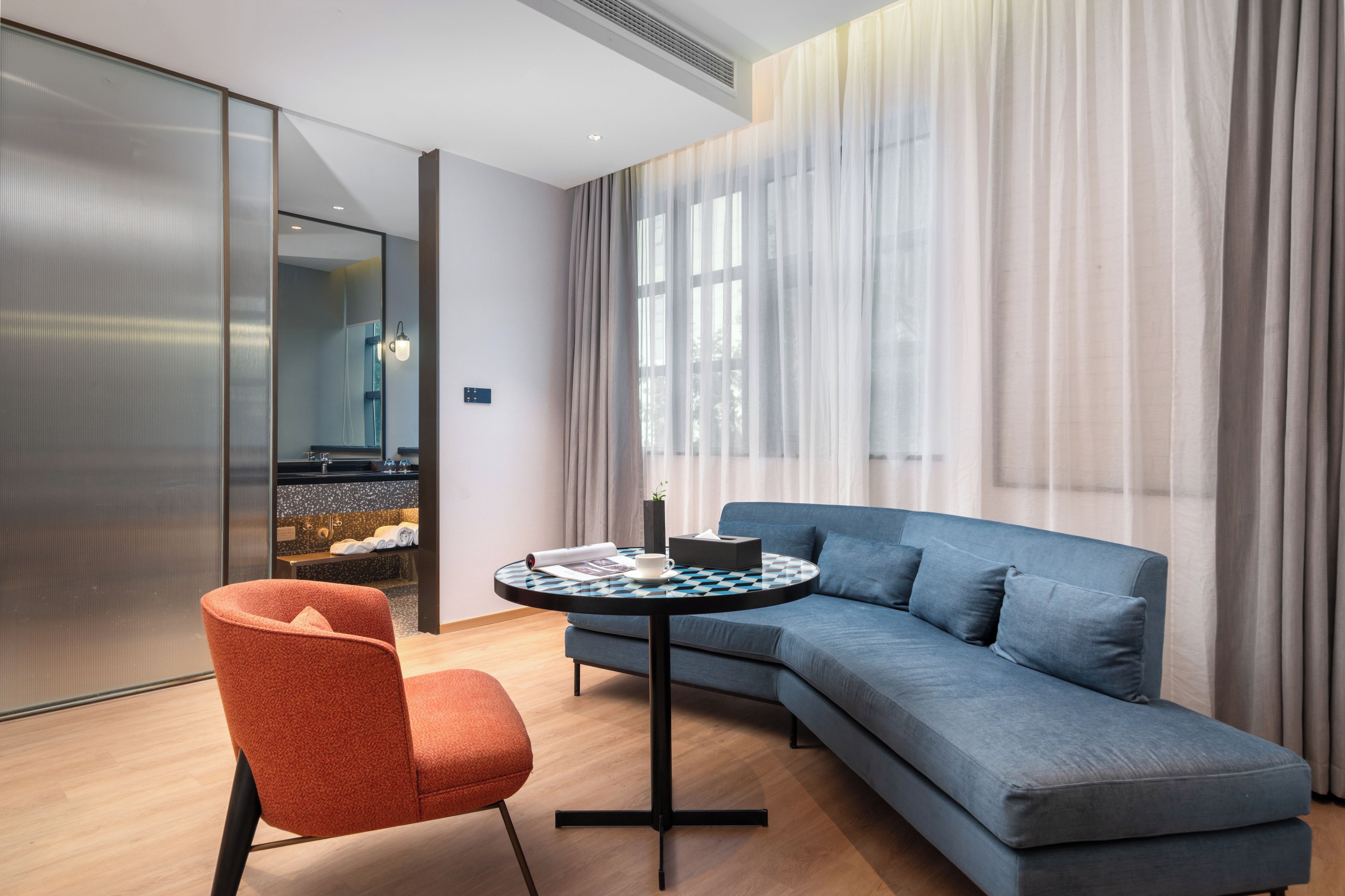 Una habitación de hotel moderna con un sofá gris, una mesa redonda y una silla roja.