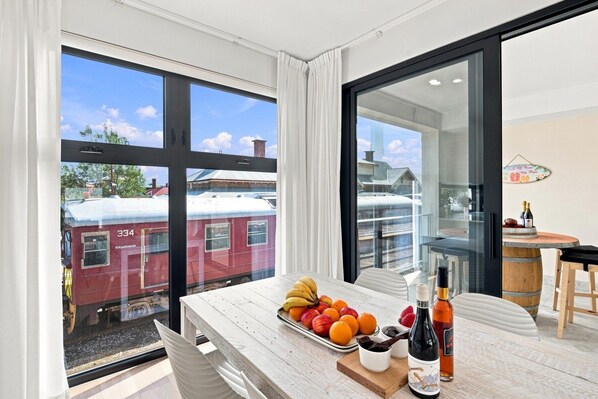 A dining area with a table set with fruits and bottles, large windows with a view of a train, and a modern interior design.