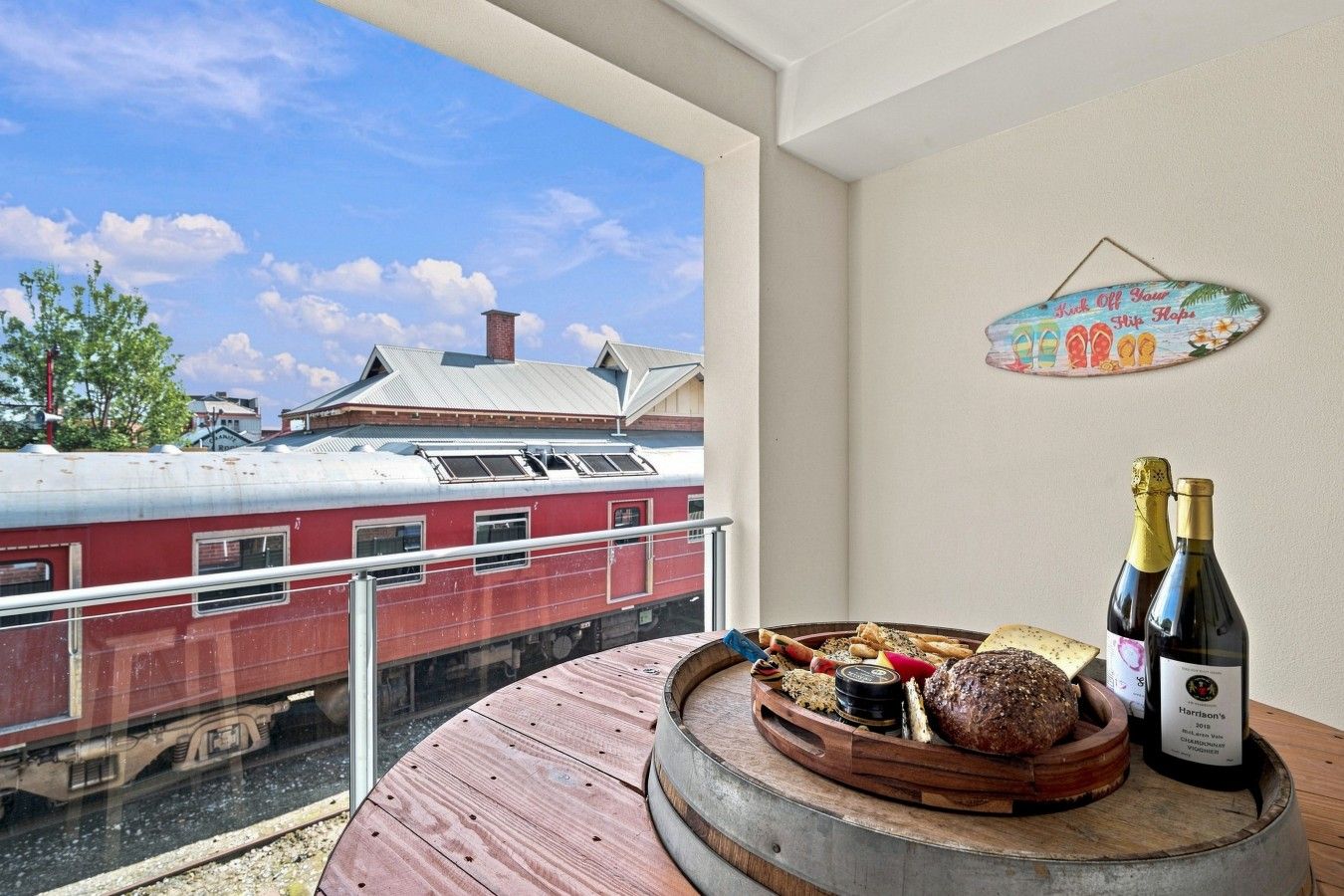 A balcony with a view of a red train, a wooden table with food and drinks, and a wall decoration.