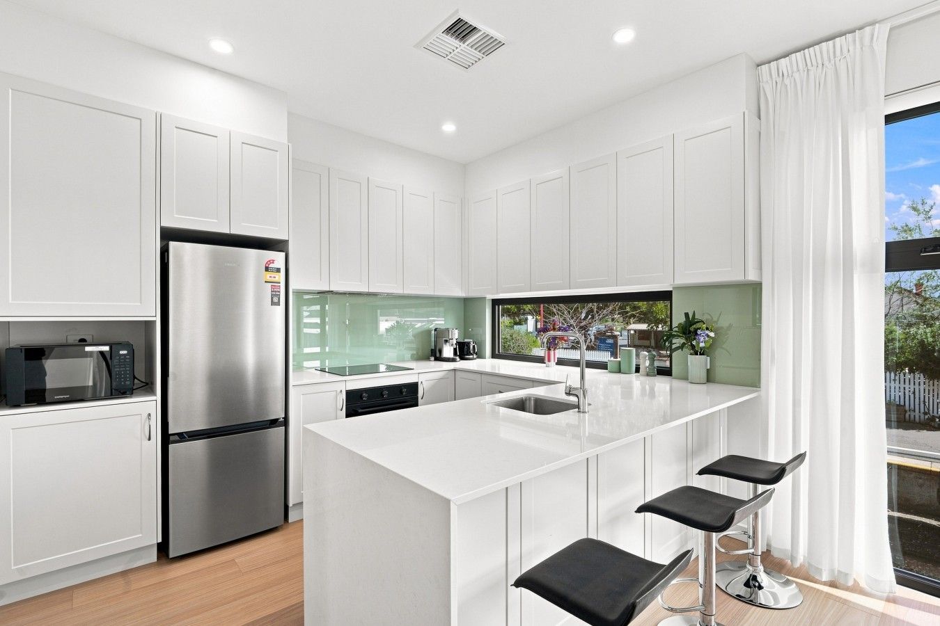 A modern kitchen with white cabinetry, stainless steel appliances, a central island with bar stools, and a large window offering a view of the outdoors.