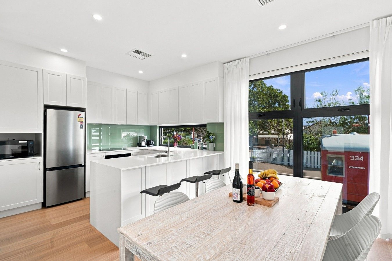 A modern kitchen with white cabinetry, a stainless steel refrigerator, a large island with bar stools, and a dining table with a fruit bowl and bottles.