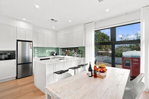 A modern kitchen with white cabinetry, a stainless steel refrigerator, a large island with bar stools, and a dining table with a fruit bowl and bottles.