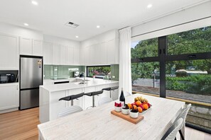 A modern kitchen with white cabinetry, a stainless steel refrigerator, a large island with bar stools, and a view of a green landscape through a large window. - Modern 2BR Townhouse | Coastal Views, Double Garage & Steps to the Beach (Victor Harbor)
