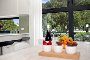 A modern kitchen with a large window, a bottle of wine, and a fruit bowl on the counter. - Modern 2BR Townhouse | Coastal Views, Double Garage & Steps to the Beach (Victor Harbor)