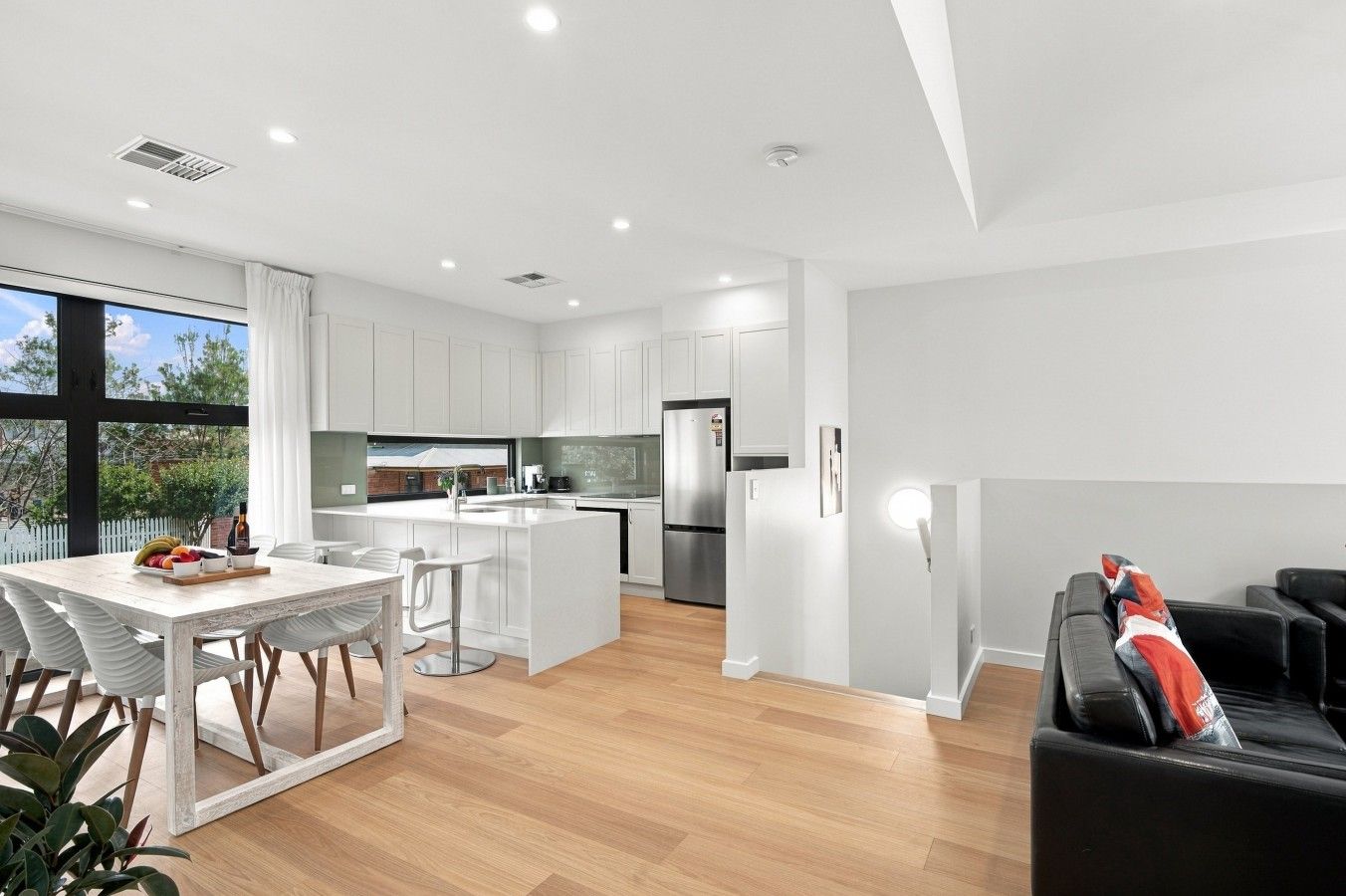 A modern kitchen with white cabinetry, a dining area with a white table and chairs, and a living area with a black sofa.