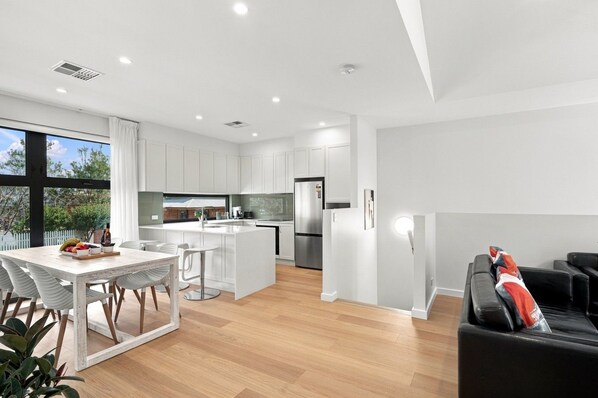 A modern kitchen with white cabinetry, a dining area with a white table and chairs, and a living area with a black sofa.