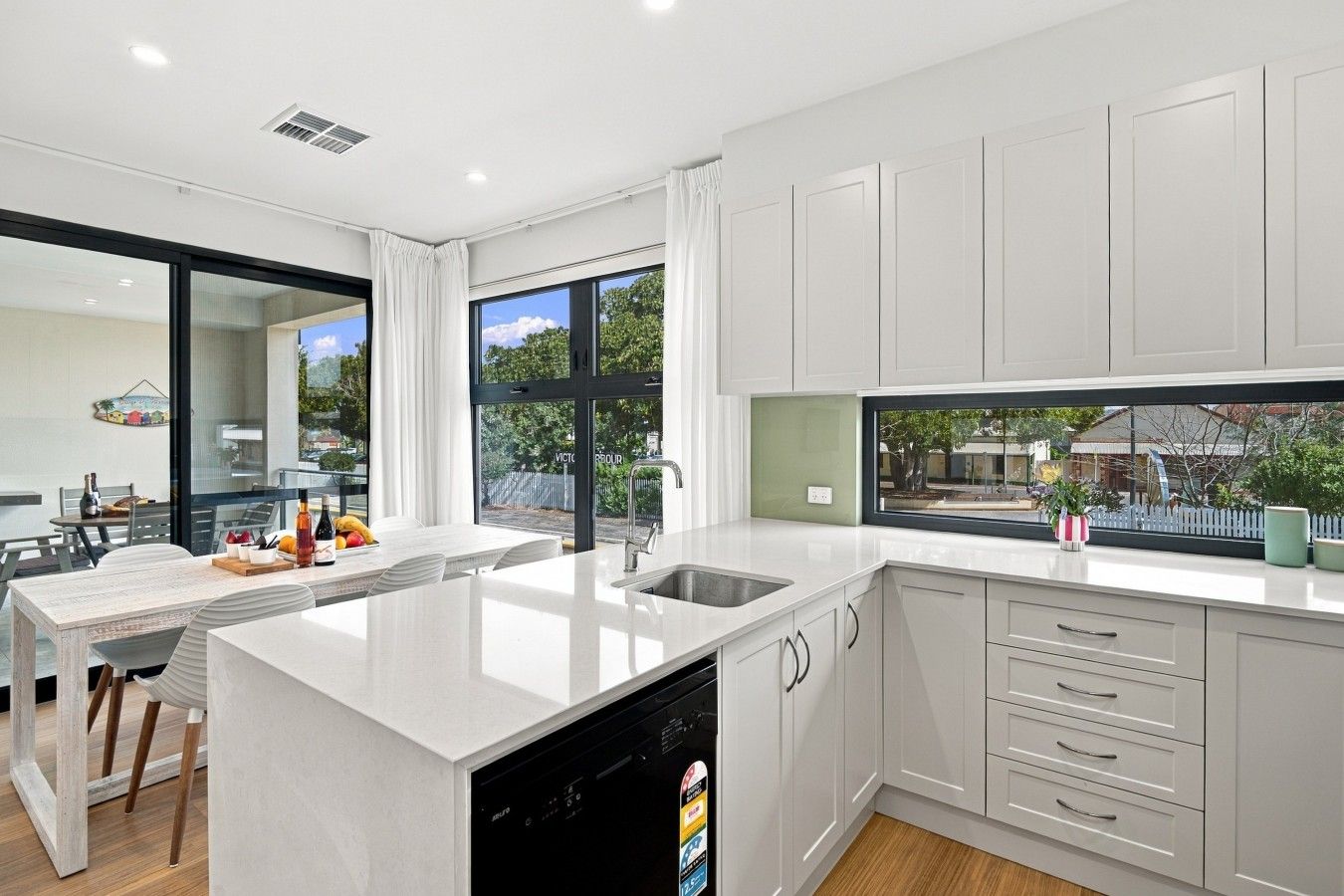 A modern kitchen with a dishwasher, a dining area with a table and chairs, and a view of the outside through large windows.