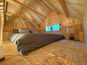 A wooden cabin bedroom with a bed, two windows, a nightstand, and a rug.