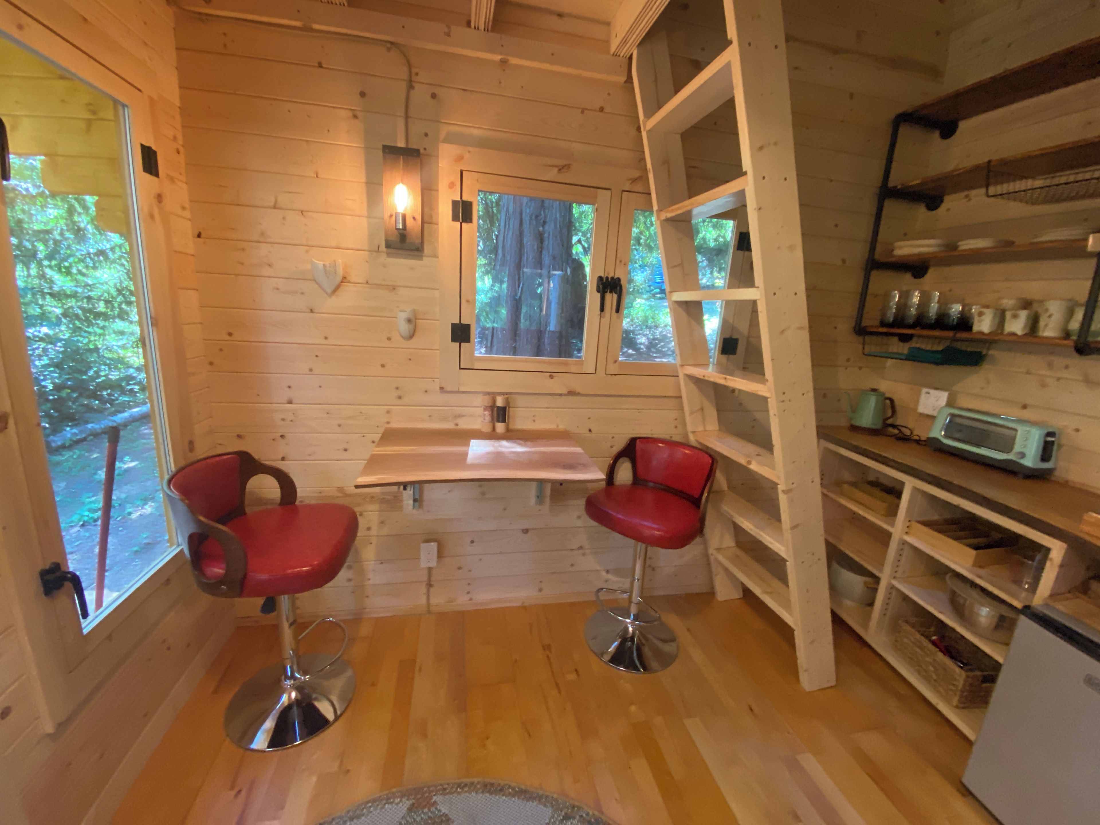 A wooden cabin interior with a dining area, two red bar stools, a window with a view of greenery, and a kitchenette with shelves.