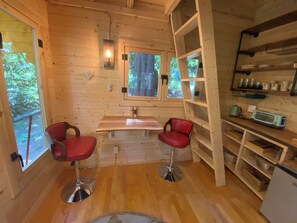 A wooden cabin interior with a dining area, two red bar stools, a window with a view of greenery, and a kitchenette with shelves. - Forest Hideout - Peaceful Cabin Getaway in the woods (Duncan)