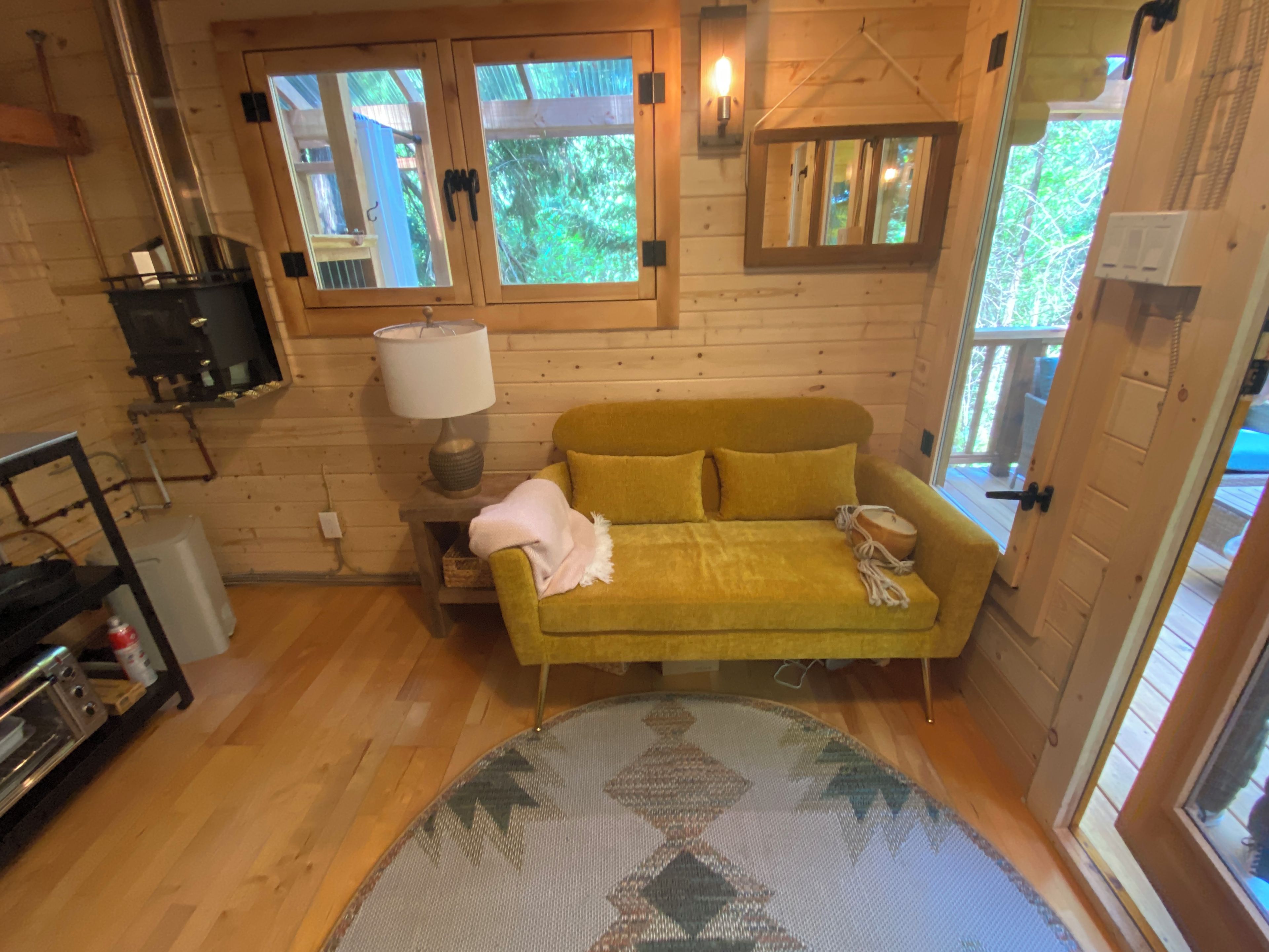 Une pièce chaleureuse avec un canapé jaune, une lampe, une petite table et une fenêtre donnant sur un paysage verdoyant.