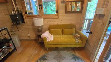 Une pièce chaleureuse avec un canapé jaune, une lampe, une petite table et une fenêtre donnant sur un paysage verdoyant.
