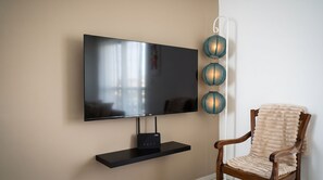 Ein flachbildfernseher an einem schwarzen Ständer an der Wand, mit einem Holzstuhl und einer flauschigen beige-farbenen Decke daneben.